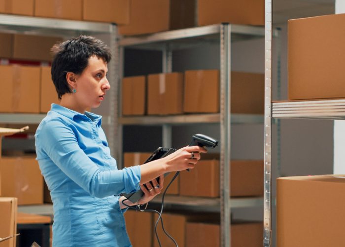 Business owner scanning stock from storehouse shelves, working with scanner to do inventory and check logistics supplies. Female employee using tablet to scan bar code on merchandise. Handheld shot.